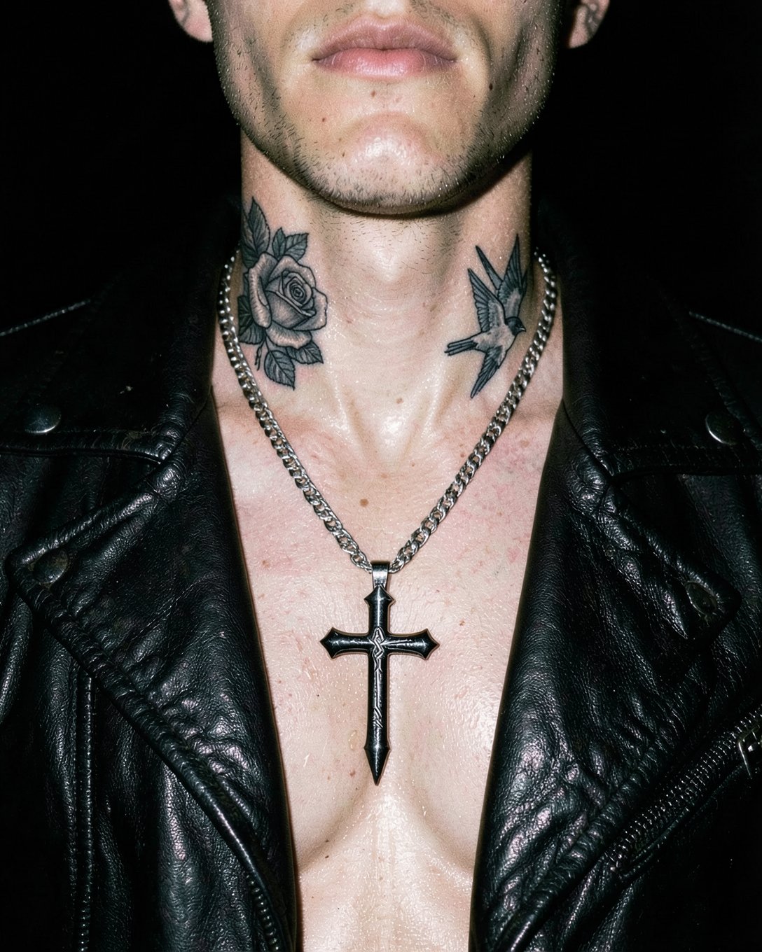 At night under a hard flash, a heavy sterling silver dagger cross pendant on a curb chain rests on the bare chest of a man in an open black leather moto jacket, a black and grey rose and swallow tattooed on his neck.