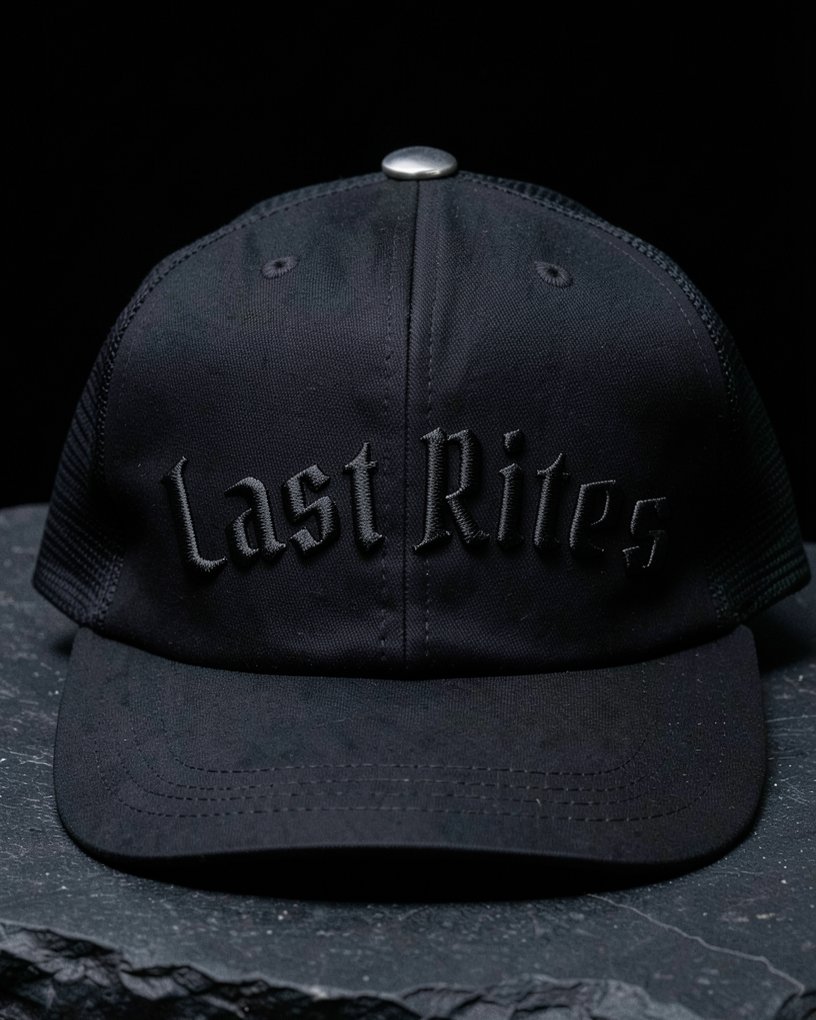 A black trucker hat with Last Rites in raised black-on-black gothic puff embroidery and a sterling silver top button.