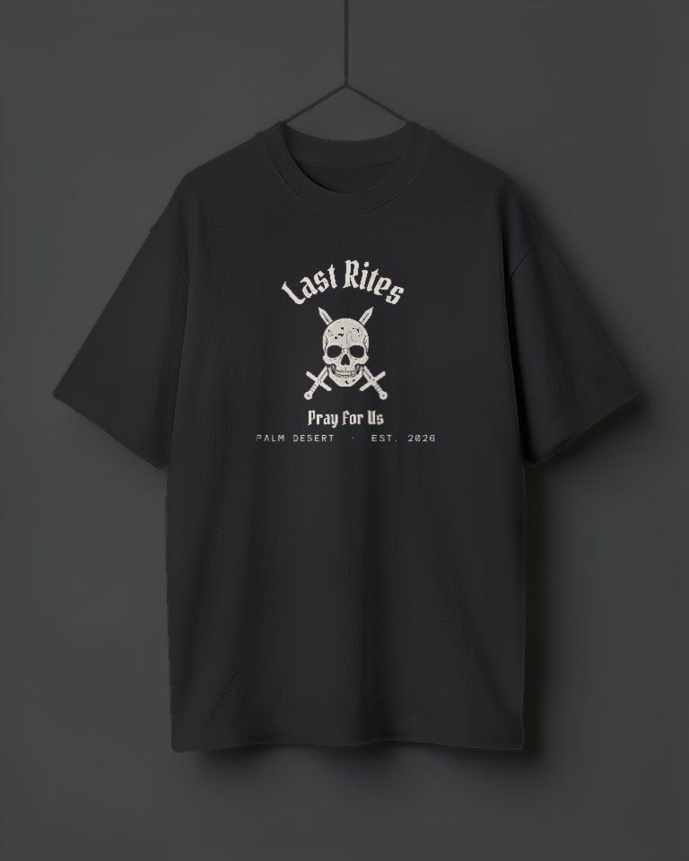 An oversized washed black tee hanging on a thread, printed in cracked cream with Last Rites arched over the skull and crossed swords, Pray For Us, and Palm Desert, est. 2026.